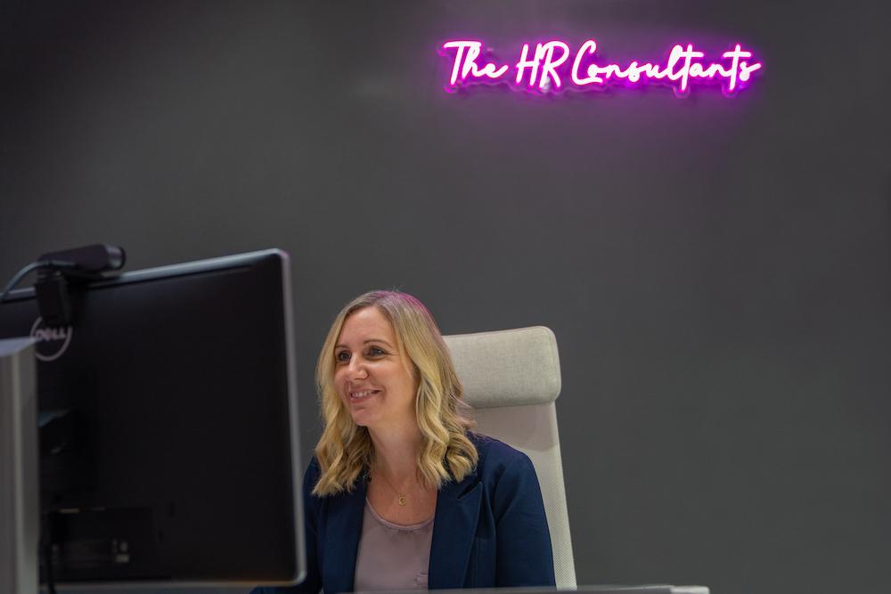 Person working at desk computer with Neon Sign behind them, reading "The HR Consultants"